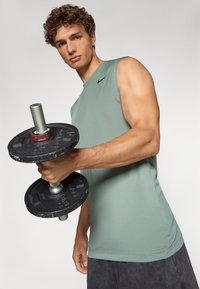 Muskel tanktop i afdæmpet grøn stof, uden ærmer. Mørk vægtløftningsdumbbell holdt i hånden, med sorte plader og sølvfarvet stang.