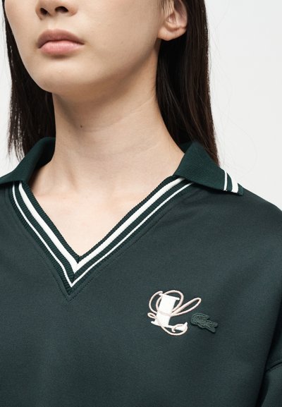 Jeune personne portant un polo vert foncé avec un col en V rayé blanc et des logos brodés L et crocodile sur la poitrine.