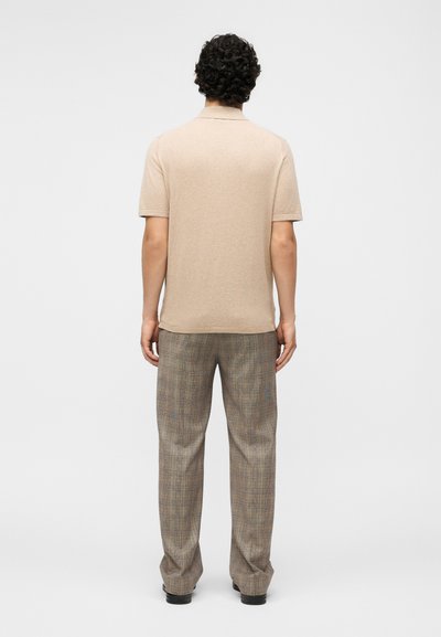 Bărbat cu păr creț, cu fața în altă direcție, purtând un tricou polo bej cu mâneci scurte și pantaloni gri cu carouri, lejeri, și pantofi negri.