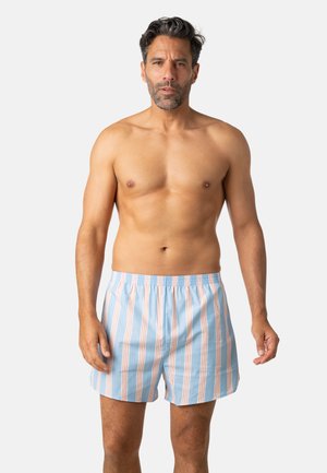 Homme à la peau mate, cheveux courts foncés, barbe, debout torse nu portant un boxer bleu clair avec des rayures verticales orange et blanches.