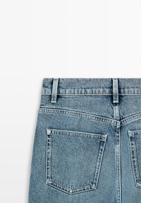 Achteraanzicht van lichtblauwe denim jeans met gestikte achterzakken en lussen voor een riem op een witte achtergrond.