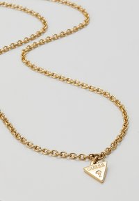 Collier chaîne en or avec un petit pendentif triangulaire gravé du mot "GUESS" et d'un symbole de point d'interrogation.