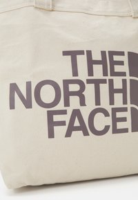 Bolsa de lona beige con un gran texto de logo marrón oscuro que dice "THE NORTH FACE". Textura suave y diseño resistente.