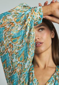 Mujer con largo cabello castaño que lleva una blusa de patrones azules y marrones, levantando su brazo para cubrir parte de su cara contra un fondo liso.