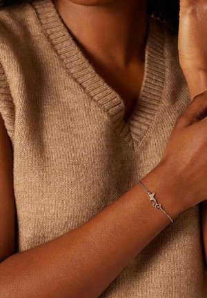 Zilveren armband met een abstract ontwerp, met een dunne ketting en een minimalistische bedel, gedragen op een gebruinde arm, tegen een beige trui.