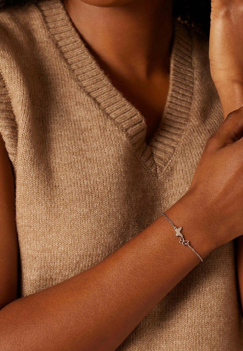 Zilveren armband met een abstract ontwerp, met een dunne ketting en een minimalistische bedel, gedragen op een gebruinde arm, tegen een beige trui.