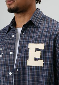 Geruit overhemd met knopen in marineblauw en bruin, met wit geborduurde tekst en een grote beige letter 'E' applicatie. Gemaakt van katoen, met een kraagontwerp.