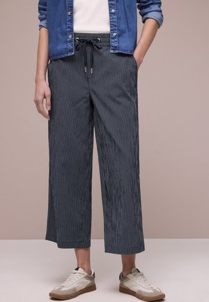 Person iført cropped, marineblå bukser med striber og snøre i taljen, hvide sneakers, en hvid skjorte og en opkørt denimjakke.