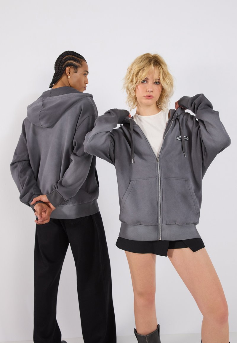 Siva zip-up dukserica s kapuljačom, sa logotipom na grudima, velikim prednjim džepovima i rebrastim manžetama. U kombinaciji sa širokim crnim hlačama i šortsevima.