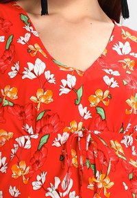 Rotes Blumenoberteil mit weißen und gelben Blumen sowie grünen Blättern; V-Ausschnitt, geraffte Taille mit einem Bindeband. Glatte Stoffqualität.