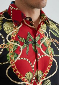 Chemise rouge avec des motifs noirs et verts, ornée de coquillages et de chaînes. Boutons en plastique, design de col classique et texture de tissu lisse.