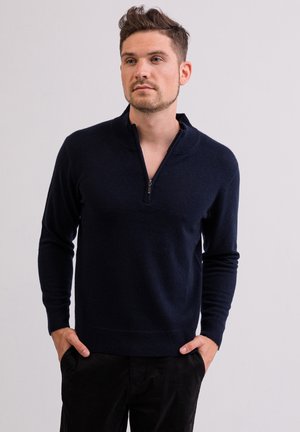 Marineblauer gestrickter Pullover mit einem Viertel-Reißverschlusskragen, gerippten Bündchen und Saum sowie einer glatten Textur. Getragen über dunklen Hosen.