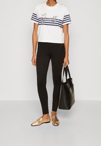 Bílý zkrácený tričko s námořnickými pruhy a grafickým textem, v kombinaci s černými legínami a metalickými mokasíny. Doplňky zahrnují černou tote tašku.