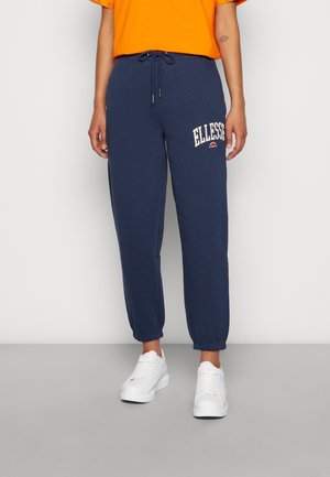 Osoba nosząca granatowe spodnie dresowe Ellesse z elastycznymi mankietami i białe sneakersy, w zestawieniu z pomarańczową koszulką z krótkim rękawem.