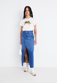 T-shirt blanc en coton avec un graphisme multicolore, jupe en jean bleu taille haute avec une fente à l'avant, associée à des baskets et un petit sac blanc.