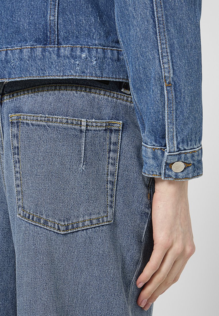 Jeansjacke in hellblauer Farbe, sichtbare Nähte und ein einzelner Knopfakzent an den Manschetten. Die Rückseite hat eine kleine Tasche.