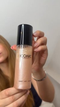 Main avec des ongles polis tenant un flacon de la base de maquillage Iconic London Prep-Set-Glow avec un bouchon noir.