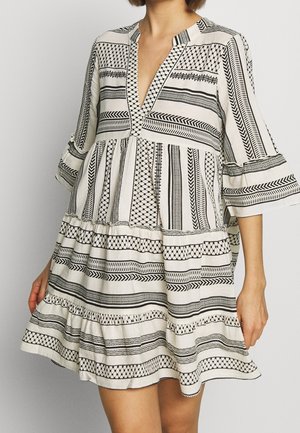 Woman wearing a loose, knee-length dress with black geometric and striped patterns on white fabric, featuring 3/4 bell sleeves and a V-neckline.
