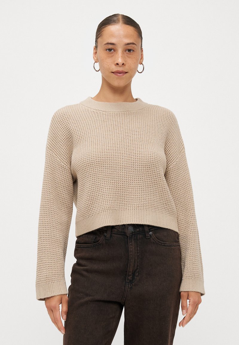 Gebreide trui in lichtbeige met een gestructureerd patroon en ronde halslijn. Verkorte lengte en lange mouwen. Gecombineerd met donkere high-waisted jeans.