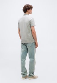 Jeune homme aux cheveux bouclés portant un polo à manches courtes gris clair, un jean bleu et des baskets beiges, debout de dos.