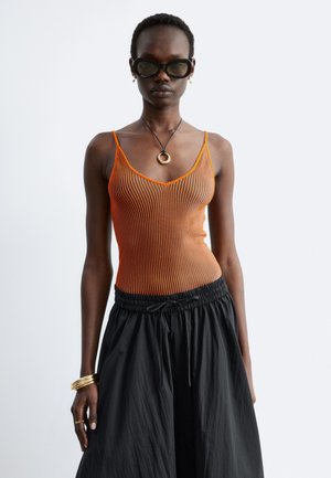 Femme portant un haut sans manches côtelé orange, un pantalon noir ample, de grandes lunettes de soleil noires, des boucles d’oreilles créoles dorées, un collier avec un pendentif circulaire et des bracelets dorés.