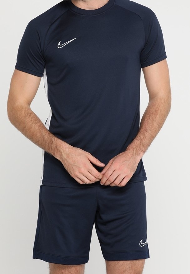 Man wearing a navy blue Nike sports t-shirt and matching shorts with white logo, standing with hands clasped at waist.
