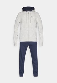 Sweat à capuche gris clair zippé avec doublure intérieure bleu marine et logo brodé, associé à un pantalon de survêtement bleu foncé avec des poignets aux chevilles.