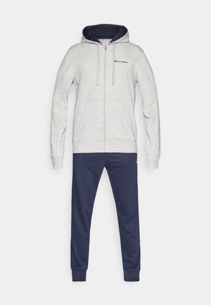 Svetlo siva jopa z zadrgo z mornarsko modro notranjo podlogo in vezenim logotipom, združena s temno modrimi športnimi hlačami z manšetami pri gležnjih.