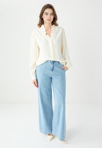 Kremowa bluzka z falbanką przy kołnierzyku, z guzikami z przodu; jasnoniebieskie szerokie jeansy; beżowe szpilki w szpic; gładki materiał, luźny krój.