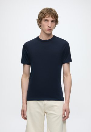 Ung mand med krøllet lyst brunt hår iført en mørkeblå T-shirt med korte ærmer og lyse beige bukser, stående foran en ensfarvet baggrund.