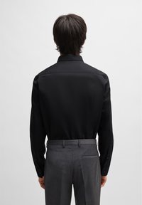 Camicia nera a maniche lunghe con colletto strutturato, texture liscia e polsini con bottone, abbinata a pantaloni grigi su misura con tasca sul retro.