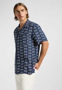 Chemise à manches courtes bleu marine avec un motif géométrique blanc, dotée d'une coupe décontractée et d'une poche poitrine, fabriquée en tissu lisse.
