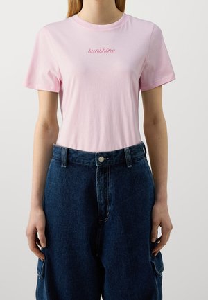 Femme portant un t-shirt rose clair avec le texte « sunshine » et un pantalon en jean bleu foncé taille haute avec des poches sur les côtés.