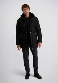 Man in een zwarte, knielange gewatteerde jas, donkere grijze broek en zwarte nette schoenen, staande tegen een effen achtergrond.