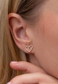 Zlatý náušničkový peříčkový tvar s minimalistickým designem, který se vyznačuje hladkými křivkami a lesklým povrchem, umístěný na bledé pleti a vlasech.