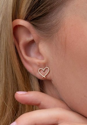 Goldene, herzförmige Ohrstecker mit einem minimalistischen Design, das durch sanfte Kurven und eine glänzende Oberfläche besticht, vor blasser Haut und Haaren.