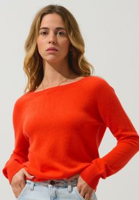 Pull orange vif en tissu doux avec un large col et des manches longues, associé à un jean en denim bleu clair. Texture lisse.