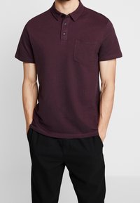 Homme portant un polo à manches courtes bordeaux foncé avec une poche sur la poitrine, associé à un pantalon noir, les mains dans les poches.