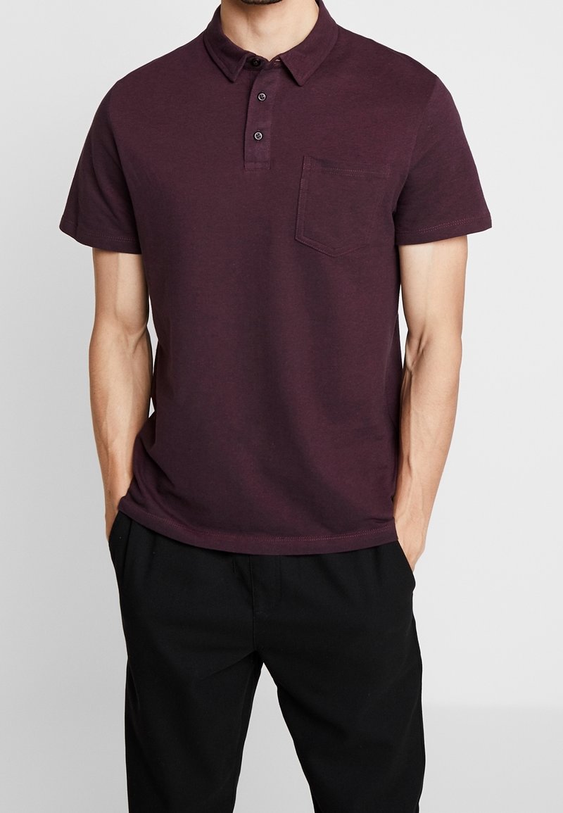 Homme portant un polo à manches courtes bordeaux foncé avec une poche sur la poitrine, associé à un pantalon noir, les mains dans les poches.