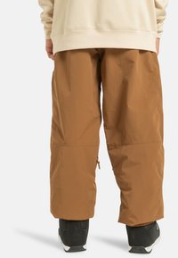 Bruine, losse buitenbroeken gemaakt van lichtgewicht materiaal, met ritsen bij de enkels, gedragen met zwarte schoenen.