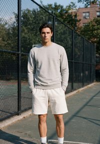Jeune homme en sweat-shirt gris clair et short blanc, debout les mains dans les poches sur un terrain de sport extérieur près d'une clôture en grillage métallique.