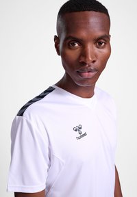 Camiseta atlética blanca con un acento negro de patrón en los hombros y un logo en el pecho, hecha de un tejido suave y ligero.