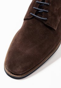 Chaussure de cérémonie en daim marron avec des lacets bleu foncé et des coutures détaillées sur le bord de la semelle, sur fond blanc.