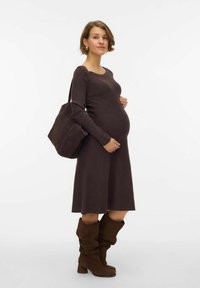 Grūtnieču kleita tumši brūnā adītā audumā, ceļgalu garuma ar garām piedurknēm, saskaņota ar brūnām samta zābakiem un atbilstošu tote somu.