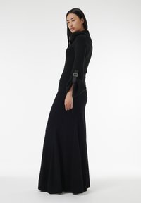Zwarte langslavige trui met een opgevouwen kraag en een gespdetail op de mouwen, gedragen met een vloeiende zwarte maxi rok.