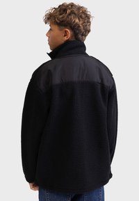 Mustat fleece-takki, jossa on teksturoitu vartalo ja sileä musta nailoninen yläselkä. Korkea kaulus ja rento istuvuus näkyvillä saumoilla.
