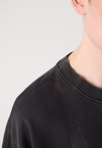 Sudadera gris oscura con un cuello acanalado. Presenta el nombre de la marca "HUGO" en relieve en el hombro. Material suave con textura mínima.