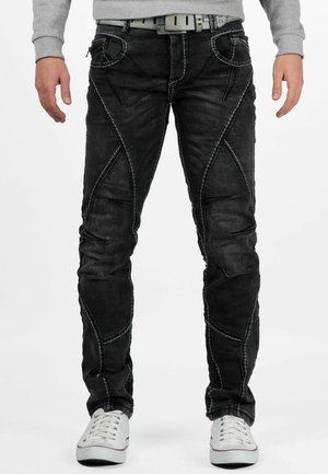 Homme portant un jean slim noir avec des détails de panneaux cousus, un sweatshirt gris, des baskets blanches et une ceinture gris clair, debout sur fond blanc.
