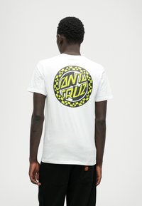 Λευκή βαμβακερή μπλούζα με κοντά μανίκια, που διαθέτει μια κυκλική γραφική παράσταση σε μαύρο και κίτρινο με το κείμενο "SANTA CRUZ" στην πλάτη. Στρογγυλή λαιμόκοψη.