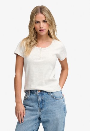 Femme blonde portant un t-shirt henley blanc à manches courtes et un jean bleu taille haute, debout avec une main dans la poche.